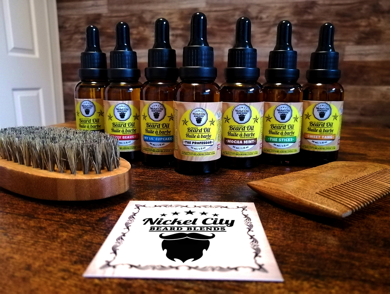 Set of beard oil bottles with a brush and comb on a wooden surface, featuring 'Nickel City Beard Blends' branding.