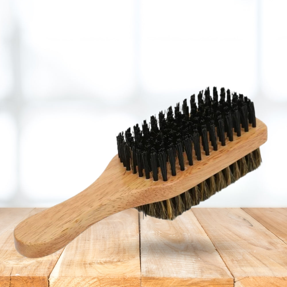 A wooden beard brush with black bristles and brown-tan natural bristles, resting on a light wood surface against a bright white background.