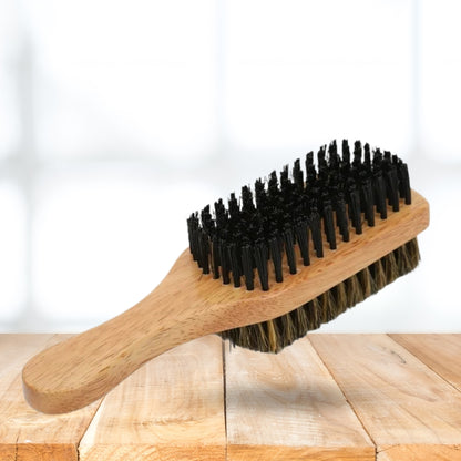 A wooden beard brush with black bristles and brown-tan natural bristles, resting on a light wood surface against a bright white background.